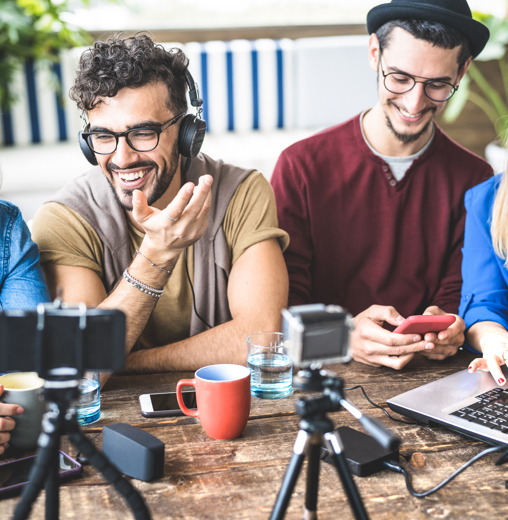 A diverse group of four young adults is gathered around a wooden table, engaged in content creation. They are smiling and laughing while interacting with various digital devices, including a laptop, smartphone, and cameras. A man with curly hair and glasses, wearing headphones, gestures expressively while speaking, while the others look on enthusiastically.